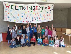 Die Kinder stehen unter einem Banner mit der Aufschrift Weltkindertag und bedruckten Händen und machen ein Peacezeichen. 