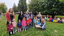 Frauen und Kinder auf einer Wiese zeigen einen Kuchen in Schlüsselform, im Hintergrund spielen weitere Kinder. 