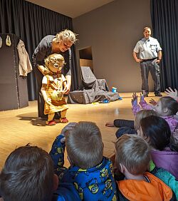 Christine Theberath steht auf der Bühne und hält eine Puppe, die das Mädchen Irmi darstellt. Mathias Reitter vom Polizeirevier Lahr steht rechts im Hintergrund ebenfalls auf der Bühne. Im Publikum sitzen Kinder, die von hinten zu sehen sind. 