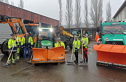 Fünf Mitarbeiter des Bau- und Gartenbetriebs Lahr in neongelben Jacken stehen mit Schneeschaufeln neben einem Räumfahrzeug.