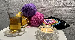 Auf dem Bild sieht man einige Wollknäule, eine Teetasse und eine brennende Kerze