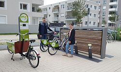 Ein junger Mann und eine junge Frau stehen an einer Mobilitätsstation mit Lastenrad und E-Bikes.