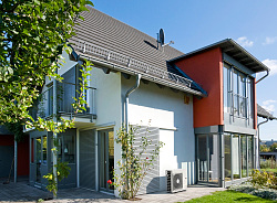 Haus vor dem ein Teil einer Wärmepumpe steht. Das Haus ist weiß und ein Bereich ist orange-rot gestrichen. es hat viele Fenster und einen Balkon.