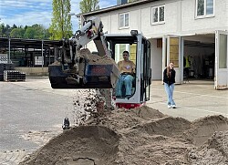 Ein Mädchen sitzt in einen kleinen Baggern und schaufelt Sand von einem Haufen. Ein weiteres Mädchen steht daneben.