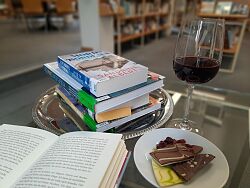 Auf dem Foto sind Bücher, Wein und Schokolade auf einem Glastisch zu sehen.