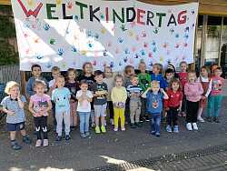 Gruppenbild der Kinder der Kita Am Schießrain vor einem großen, bunten selbst gemalten Plakat auf dem steht &#34;Weltkindertag&#34;.