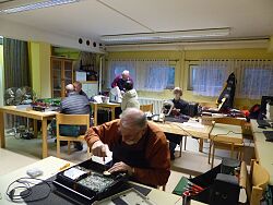 In einem Raum sitzen an verschiedenen Tischen Menschen, die Dinge reparieren.