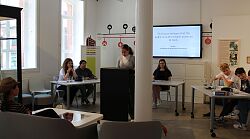 Eine Schülerin steht an einem Rednerpult, weitere Schülerinnen und Schüler sitzen an Tischen oder auf Sesseln