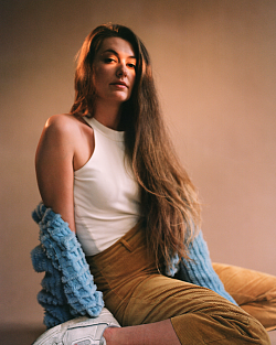 Eine Junge Frau mit langen braunen Haaren und einem weißen ärmellosen Oberteil.