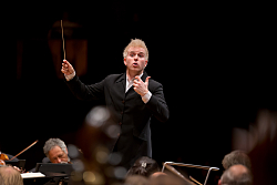 Ein Dirigent mit hellen Haaren beim dirigieren eines Orchesters.