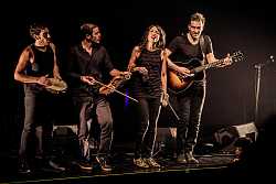 Eine Band, alle sind schwarz gekleidet. In der Mitte steht eine Frau die singt um, sie herum drei Männer jeder spielt ein Instrument,