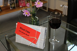 Das Foto zeigt ein in Zeitungspapier verpacktes Buch, ein kleines Blumensträußchen und ein Glas Rotwein auf einem Tisch