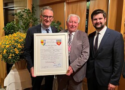 Drei Männer  in Anzügen stehen zusammen, der eine Mann überreicht dem anderen eine Würdigung die in einem weißen Bilderrahmen ist.