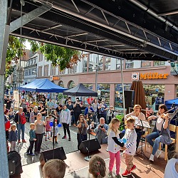 Zwei Kinder stehen auf einer Bühne, halten sich an den Händen und drehen sich umeinander im Kreis. Zwei Musikerinnen mit Instrumenten sind ebenfalls auf der Bühne. Vor der Bühne stehen Zuschauer.