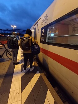 Junge Fahrgäste mit Fahrrad steigen in einen ICE ein, im Hintergrund blauer Abendhimel und eine Brücke.