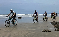 Das Foto zeigt sechs Personen die auf ihrem Mountain Bike vor den Wellen des Meeres am Strand entlang fahren.