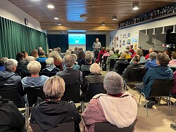 Sitzendes Publikum, von hinten fotografiert; vorne steht der Referent vor einer Leinwand