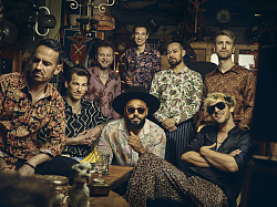 Die acht Musiker der Band FATCAT sitzen in schrille Hemden gekleidet um einen Holztisch.