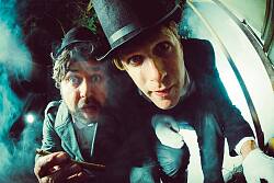 Szene aus dem Film mit Kreidlinger und Bäuerle die altmodisch gekleidet mit Hut und Zigarre in die Kamera schauen. 