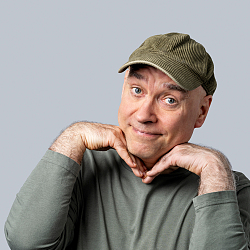 Portrait von E HG Butzko, er stützt seinen Kopf auf den Händen ab und grinst in die Kamera