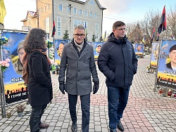 Oberbürgermeister Ibert geht mit Bürgermeister Naidaa durch die Galerie der Helden. Die gefallenen Soldaten sind mit einem Porträt auf großen Bannern aufgedruckt. Ihre Porträts zieren Fahnen in gelb-blau und rot-schwarz. 