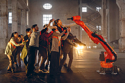 Das Foto zeigt neun Menschen in einem großen Altbau die einen weiteren Menschen hochheben und einem Roboterarm gegenüber halten.