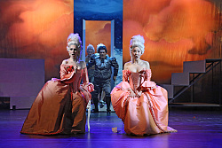 Das Foto zeigt zwei Frauen mit weißer Perücke und rosafarbenem Ballkleid auf der Bühne. Hinter ihnen tritt ein Mann mit grauer Perücke, grauer Kleidung und weißem Gesicht hervor. Ihm folgen zwei weitere Männer mit weißer Perücke und ebenfalls weißem Gesicht. Der Bühnenhintergrund stellt orangefarbene Wolken dar und in der Mitte ist einer Art Tor zu erkennen.