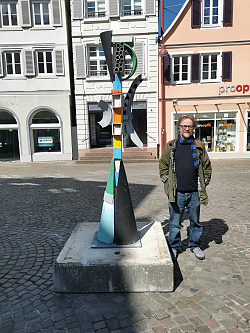 Das Foto zeigt den Künstler Menno Fahl neben seiner Skulptur in der Lahrer Innenstadt. Die Skulptur besteht teilweise aus Holz und ist verschiedenfarben bemalt. Sie steht auf dem Lahrer Sonnenplatz.