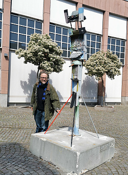 Das Foto zeigt den Künstler Menno Fahl mit seiner Skulptur &#34;Stangenfigur&#34;. Diese hat eine große Stange als Fuß an dem weitere hölzerne Stücke befestigt sind, sowie Metallgitter und eine Art Hut als oberste Spitze der Figur. Sie ist graufarben und hat blaue, gelbe und grüne Akzente.