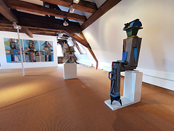 Das Foto zeigt die Skulptur des Künstlers Menno Fahl im Ausstellungsraum der städtischen Galerie.  Diese Skulptur besteht aus verschiedenen Holzstücken und ist mit Metallstücken verziert. Die Skulptur sitzt auf einem weißen Podest und hat verschiedene buntfarbige Akzente.