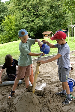 Kinder spielen im Sandkasten. Gemeinsam halten sie ein Rohr durch das Wasser gespült wird.
