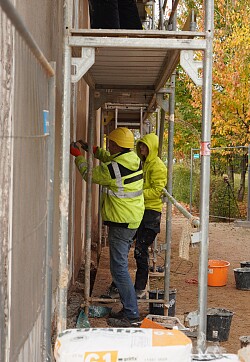 FSJlerinnen und FSJler der Jugendbauhütte Baden-Württemberg bringen an der Nordwand des Streifenhauses der Lahrer Römeranlage Kalkputz auf. die Person auf dem Bild trägt eine neongrüne Jacke.