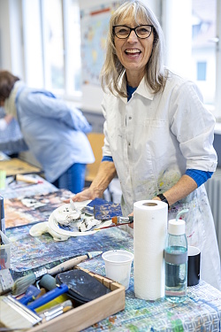 VHS-Programm der VHS Lahr bietet auch Acrylmalkurse - Person in weißem Malkittel steht an einem mit Farbklecksen bedeckten Tisch mit Malutensilien und Papierrolle, im Hintergrund eine weitere Person in blauer Kleidung.