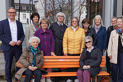 Vertreterinnen des Frauenpolitischen Forums und von Inner Wheel mit Oberbürgermeister Markus Ibert (links) und Fabian Roßmanith (Abteilung Grün und Umwelt) präsentieren die Orangene Bank. Sie stehen um die Bank gruppiert. 