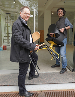 Wirtschaftsförderer Robin Derdau (Stadt Lahr) und Dr. Carsten Hutt (Werk ISI, Reutlingen) richten die Werk.Stadt.Lahr in einem Leerstand am Lahrer Schlossplatz ein. Sie beide stehen an der  Eingangstür und tragen jeweils einen Stuhl in das Innere des Leerstands. 