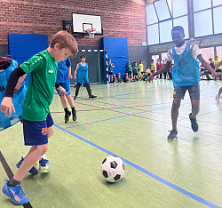 Auf dem Bild sind Kinder in sportlicher Bekleidung zusehen die in einem Turnier mitspielen. Sie spielen in einer Turnhalle. Sie tragen verschieden Team Trikots. 
