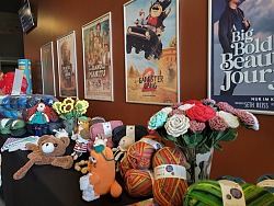 Auf dem Bild sind verschiedene Wolle arten und Farben zusehen, welche auf einem Tisch liegen. Auf dem Tisch liegen auch verschiedene gestrickte Gegenstände, wie Plüschtiere und Blumen. Dahinter befinden sich Kinoplakate an der Wand.