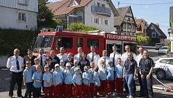 Kindergruppe in blauen T-Shirts und roten Hosen steht vor einem roten Feuerwehrfahrzeug mit mehreren Erwachsenen in Uniformen auf einer Straße in einem Wohngebiet