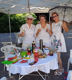 Drei Frauen in weißer Kleidung stehen unter einem Pavillon an einem Tisch mit verschiedenen Speisen und Getränken, eine Frau hält einen weißen Sonnenschirm