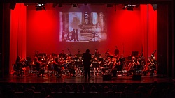 Orchester mit Streichern und Bläsern auf Bühne vor rotem Hintergrund und projiziertem Schwarz-weiß-Film