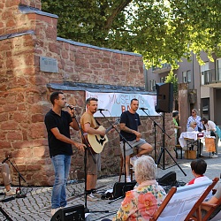 Musiker spielen vor einer alten Steinmauer auf einem gepflasterten Platz vor Publikum, das in Liegestühlen sitzt
