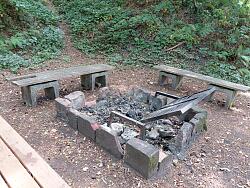 Feuerstelle mit verkohltem Holz und beschädigtem Grillrost umgeben von drei einfachen Holzbänken in einem Waldgebiet.