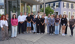 Gruppe von jungen Erwachsenen und zwei älteren Personen steht vor einem modernen Gebäude mit Glasfassade und Pflastersteinen.