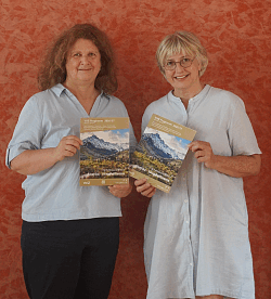 Zwei Frauen vor roter Wand halten jeweils ein Programmheft mit Berglandschaft auf dem Cover
