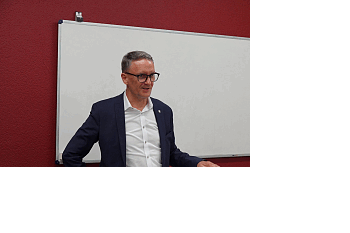 Mann mit Brille und dunklem Anzug steht vor weißem Whiteboard an Stehtisch mit Unterlagen und Smartphone