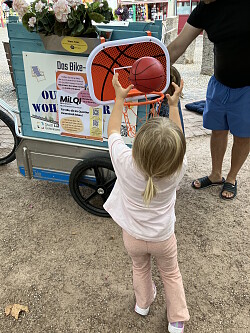 Ein kleines Mädchen wirft einen Basketball in den Basketballkorb, der an der Spielekiste befestigt ist. 