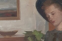 Zu sehen ist eine junge Frau mit Hut. Sie sitzt an einem Tisch und liest in einem dünnen Buch. Vor ihr auf dem Tisch steht ein kleiner Krug. Im Hintergrund ist ein Zimmer zu sehen mit Kommode, Bild und Pflanze 