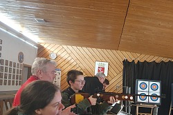 Zu sehen ist eine junge Frau, die mit einem Luftgewehr in die Ferne zielt. Sie befindet sich in einem Schießstand. Im Hintergrund sind weitere Personen zu sehen.