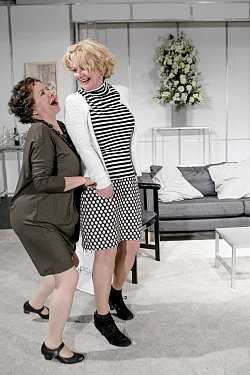 Zwei Frauen mittleren Alters, beide tragen knielange Kleider, lachen miteinander. 