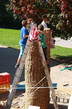 Das Bild zeigt die Klötzchenbaustelle in einer Sandkiste. Vorn stehen zwei Jungen auf einer Leiter, um an die Spitze des runden Klötzchenturms heranzukommen. Hinter dem Turm steht ein Junge, vermutlich ebenfalls auf einer Leiter, die jedoch vom Klötzchenturm verdeckt ist, sodass man nur seinen Kopf und seinen Oberkörper sieht.
